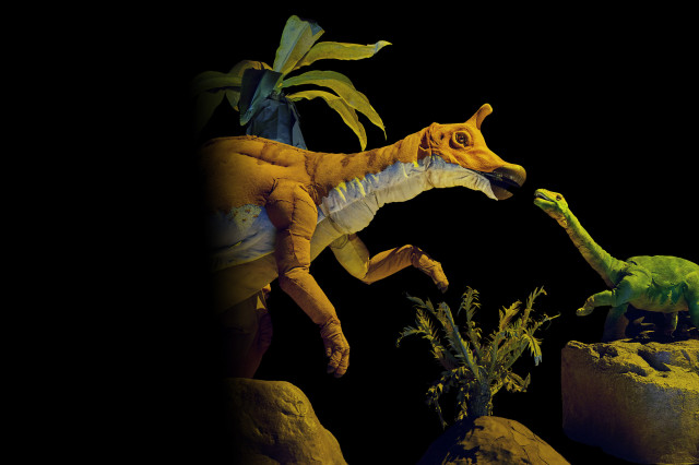 Hadrosaur orange and white puppet facing a green miniature long-necked dinosaur puppet