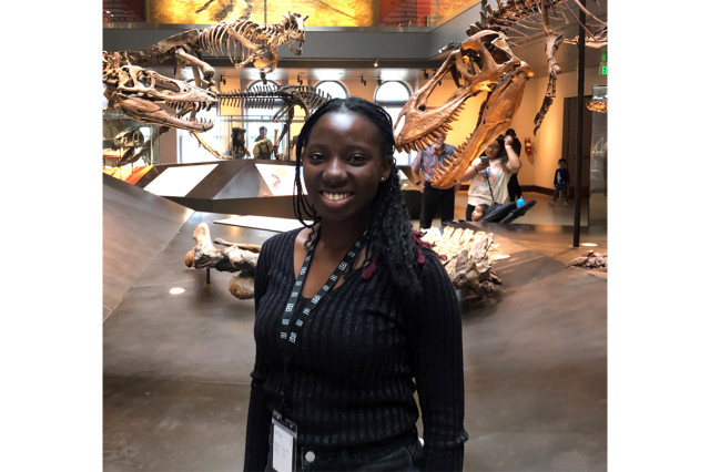 Ayomi Fadaka from the waist up, wearing a black long-sleeved shirt, with dinosaur skeletons in the background