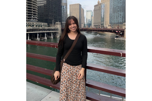 Christina Ela wearing a black sweater and flowered skirt with water and buildings in the background
