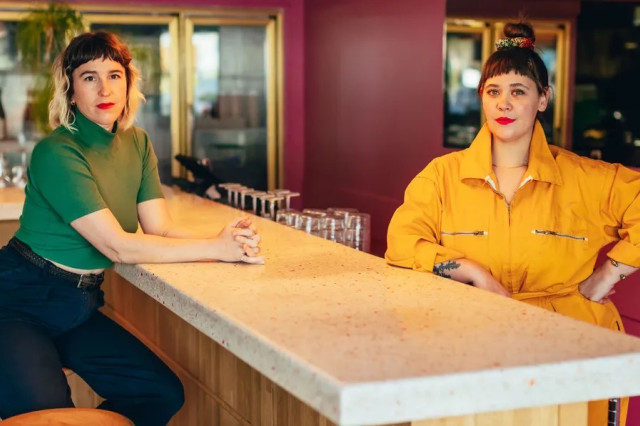 Two women posing on either side of a marble bar, one sitting and one standing.