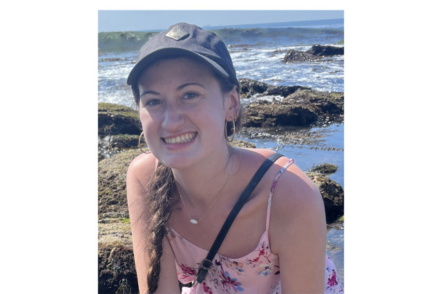 Head and shoulders portrait of Julia Morales wearing a navy baseball cap and pink tank top with rocky terrain and ocean in the background