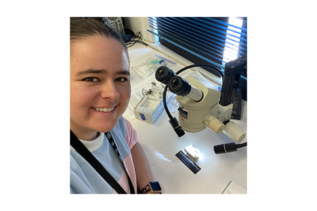 Kristin Watmore wearing a black lanyard and leaning on a table with a microscope