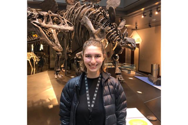 Libby Konjoyan from the waist up wearing a black shirt and puffy coat with dinosaur skeleton in the background