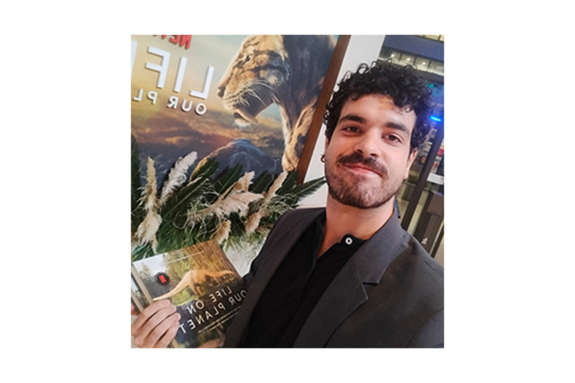 Head and shoulders portrait of Dr. Sergio Martínez Nebreda holding a book in front of a poster of a saber-toothed cat