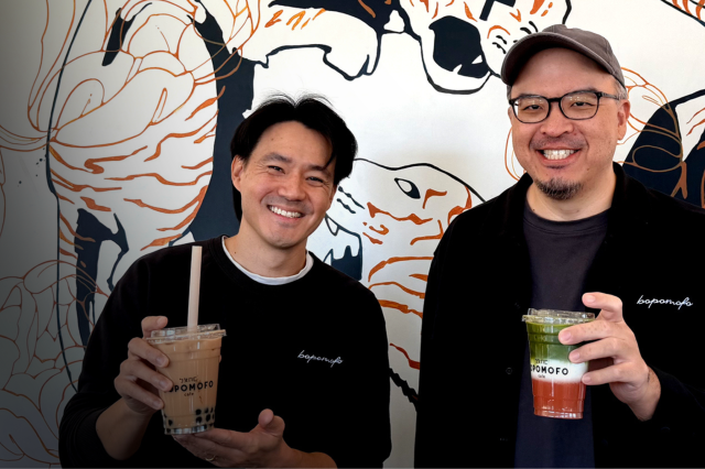 two men holding drinks, wearing black shirts, man on right wearing glasses ands a hat. Standing against graphic wall. 