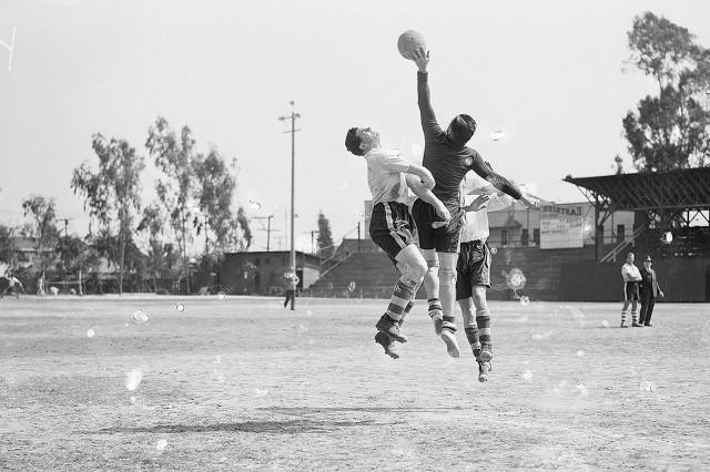 Black and white photo of three soccer players jumping in the air while one player reaches for the ball with his hand