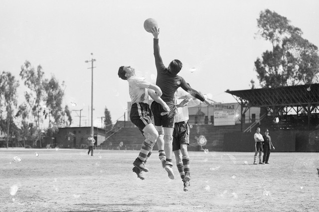 Black and white photo of three soccer players jumping in the air while one player reaches for the ball with his hand