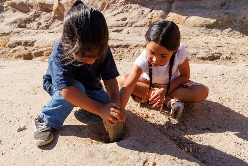 Two children from the Fernandeno Tataviam Band of Mission Indians are grinding acorns using a pestle. The boy is crouched over with both hands on the pestle and the girl is seated nearby with one hand on the pestle guiding him. 