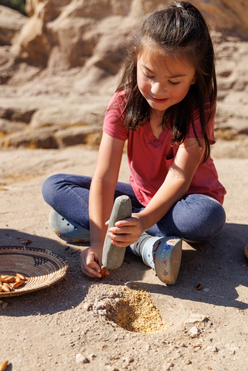 A girl from the Fernandeno Tataviam Band of Mission Indians is seated atop a bedrock mortar. She is holding an acorn upright in one hand and a pestle in the other. She looks poised to crack the acorn with the pestle. 