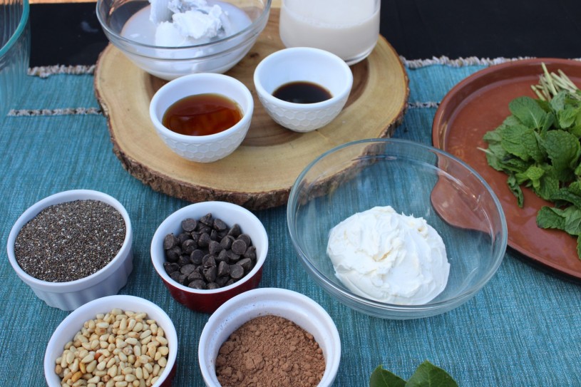 Small bowls of ingredients on a table, including cream cheese, cacao, chocolate chips, chia, milk, pine nuts, mint, and honey