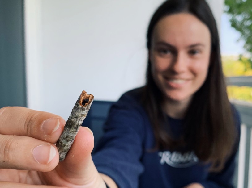 Woman out of focus holds up a twig with a tiny ant head poking out, with her hand and the twig in focus