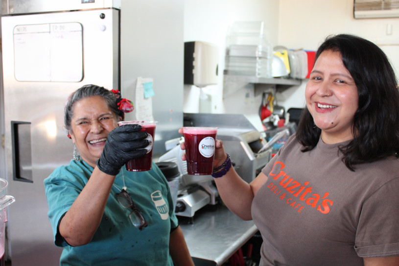 Two smiling women in a commercial kitchen holding up clear cups of dark red juice. One woman wears a brown t-shirt with "Cruzita’s Deli & Cafe" branding.