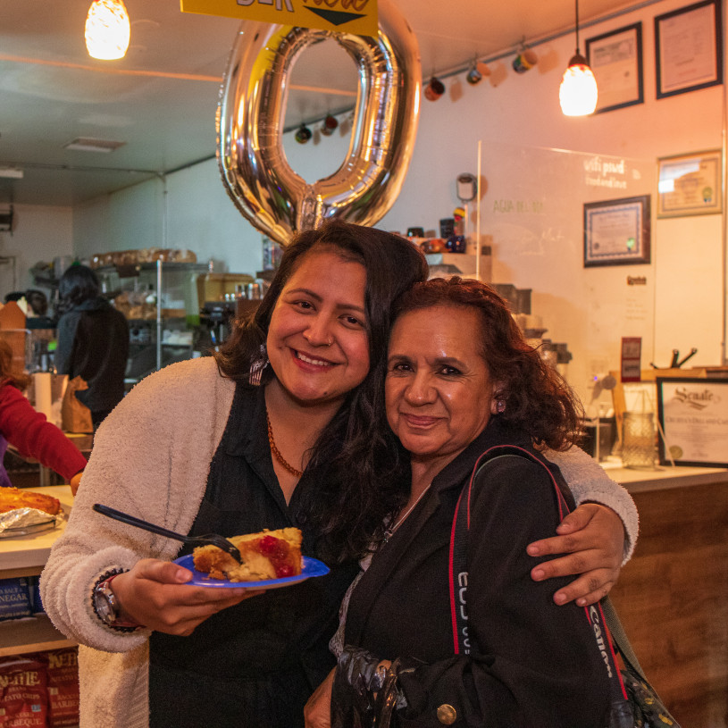 In front of a cafe counter, a young Latina woman holds a slide of cake on a plate in one hand and side-hugs her older aunt with the other.