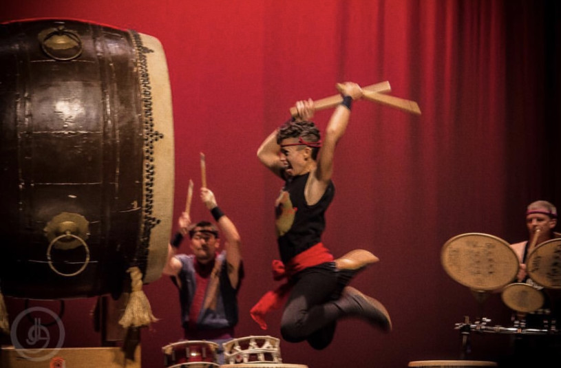 A taiko player caught mid-air and mid-strike of a big drum with drummers in the background.