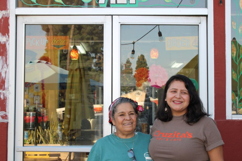 Latina mother and daughter stand below their business sign "Cruzita's Deli & Cafe".
