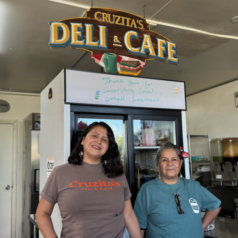 Latina mother and daughter stand below their business sign "Cruzita's Deli & Cafe".
