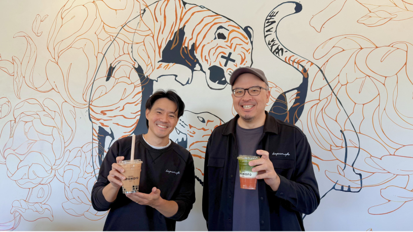 Two adults stand side by side indoors, smiling at the camera while holding iced drinks in clear plastic cups. One drink is a milk tea with tapioca pearls and a straw; the other is a layered beverage with green and reddish tones. They are positioned in front of a wall mural with abstract line drawings.