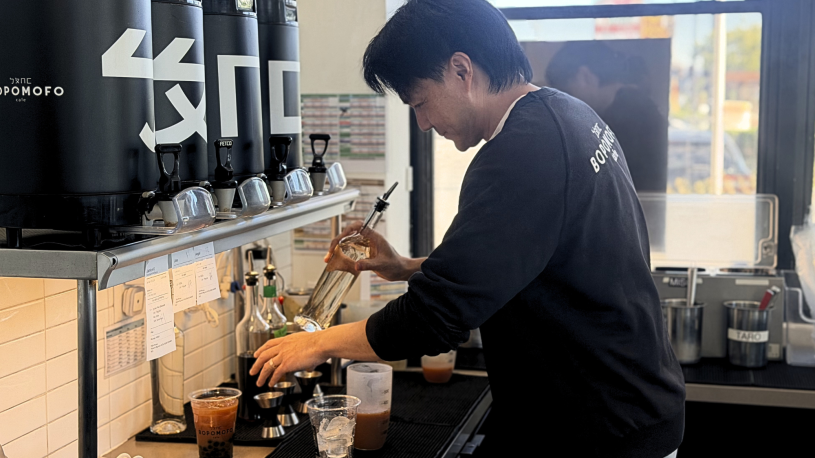 A person stands at a café drink station, pouring a brown liquid from a container into a clear plastic cup filled with ice, with syrup bottles, measuring tools, and beverage dispensers arranged on the counter nearby.