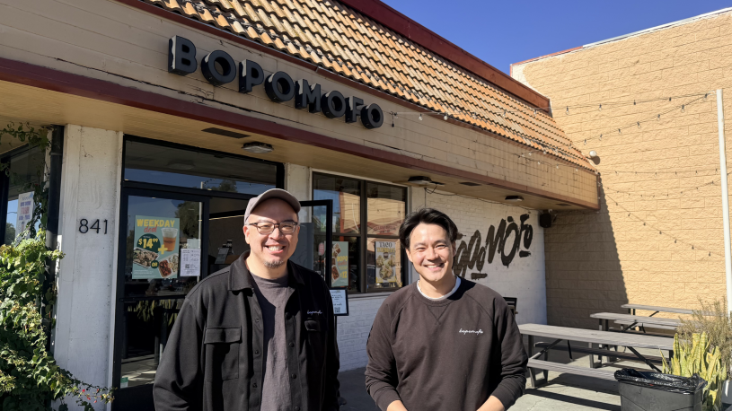Two men standing outside store front, one in glasses and hat and other in sweatshirt