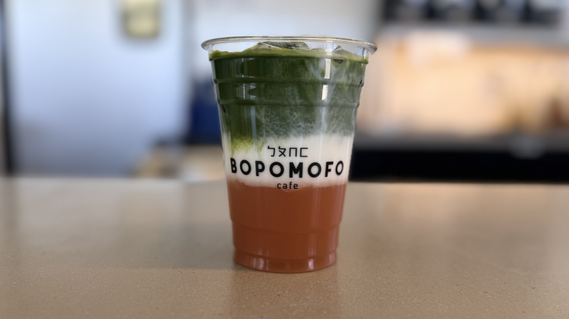 A person stands behind a café counter, pouring a green liquid from a small container into a clear plastic cup with ice, creating a layered drink with green, white, and reddish tones. A menu board and beverage dispensers are visible in the background.