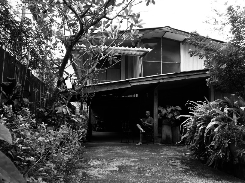 A man sits barefoot in front of his traditional Thai house reading a book.