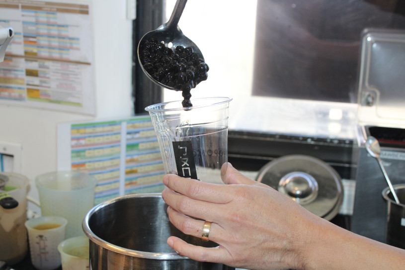 lose-up of a hand holding a clear plastic cup while a spoonful of black tapioca pearls is being added from above, with a metal pot and café prep area visible in the background.