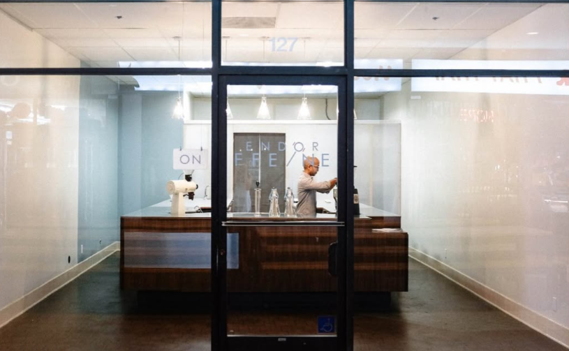 Looking through a coffee shop window from the street; inside, a single man stands behind the counter making coffee.