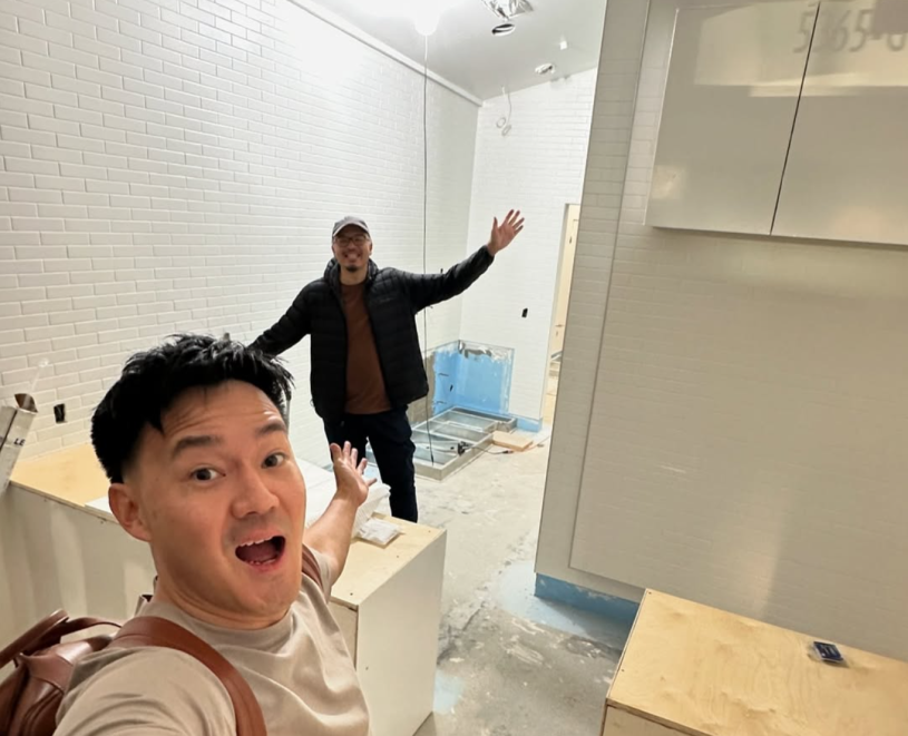 Two people stand inside a small, unfinished interior space with white tiled walls, exposed flooring, and visible construction areas. One person in the foreground takes a selfie with an excited expression while gesturing toward the room, and another stands farther back with arms raised, smiling.