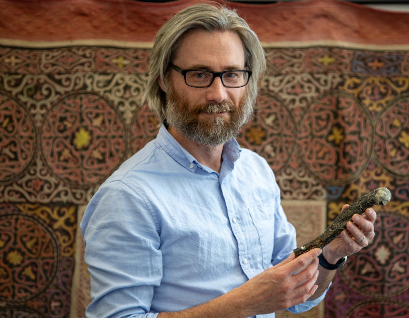 A man with long gray hair, beard, wearing glasses, in a blue collared shirt holds a fossil