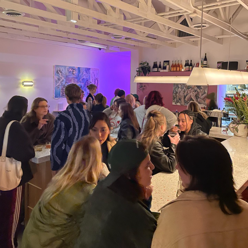 Mix of people at a small restaurant with high ceilings, some people sitting and others standing around long, white marble bar tables.