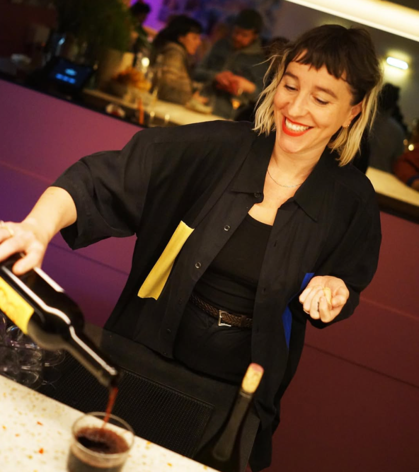 Female bartender dressed in back, pouring a glass of red wine behind a mirrored bar.