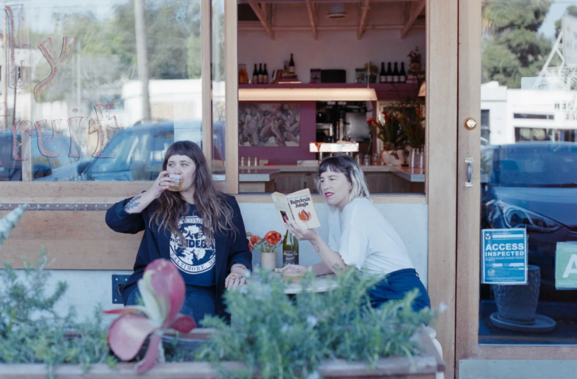 Two women sitting in outside seating in front of a restaurant; one sipping from a glass and one reading a book.
