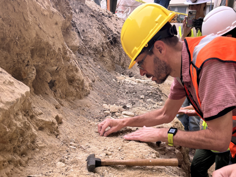 A man in a yellow helmet and orange safety vest examines dirt.