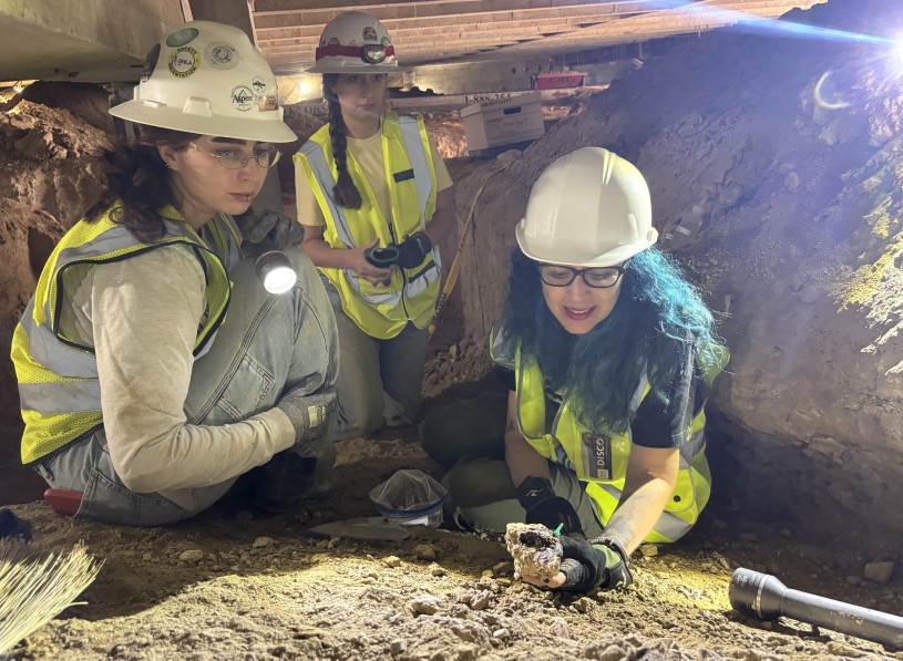 Three women in white safety helmets and yellow vests crouch over dirt.