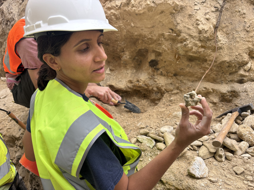 A woman in a white helmet and yellow safety vest holds up an invertebrate fossil.
