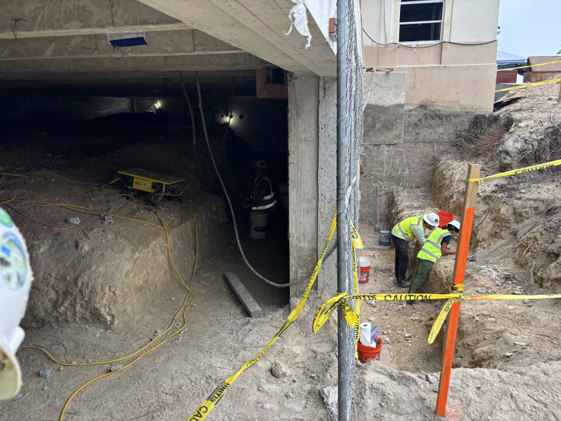 A chain link fence bisects the image with a trench on the right and construction beneath a building on the left.