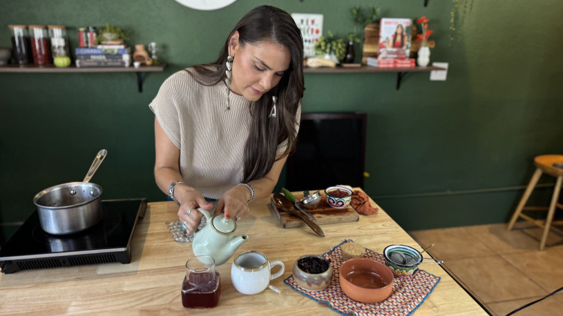 Woman pouring hot tea from a tea pot into a small cup, with the fresh ingredients used to make the tea sitting next to the cup on the counter.