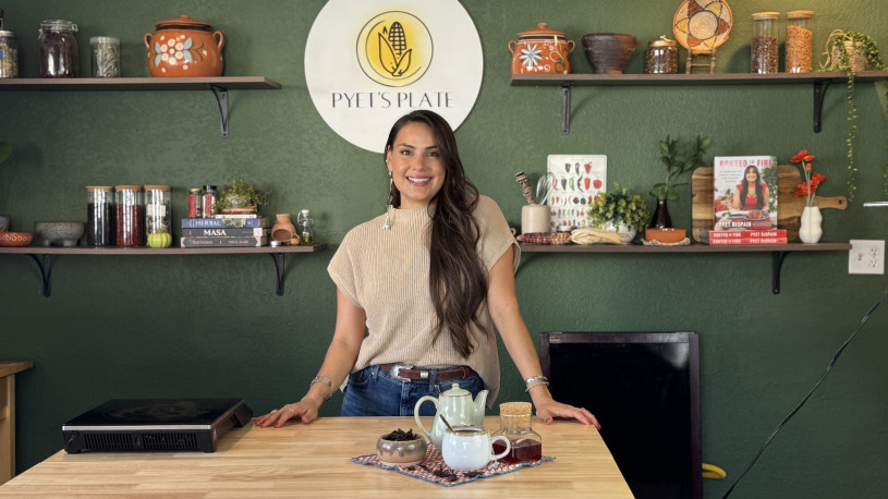 Female chef posing behind a wooden counter with tea-making ingredients on the counter and jars of ingredients and cookbooks on the shelves behind her.