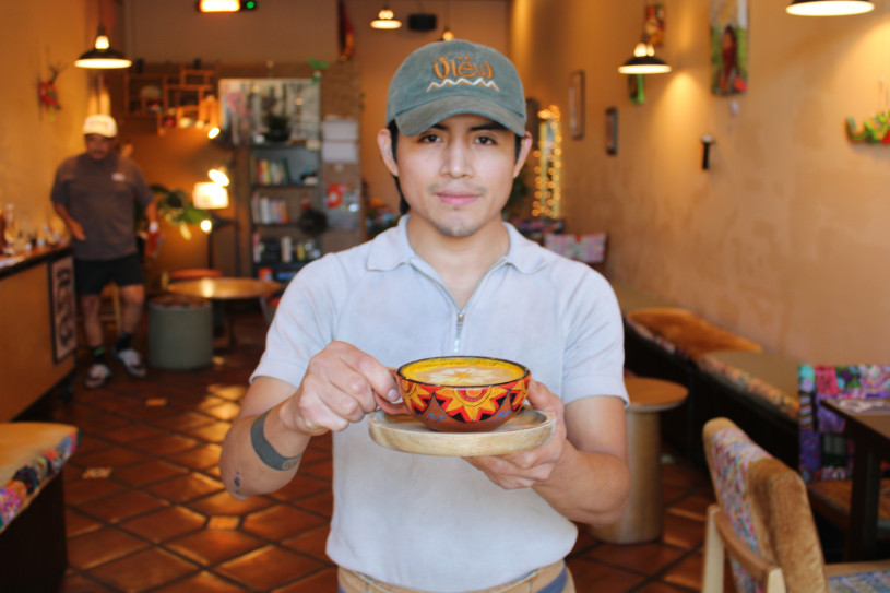 A Guatemalan man wearing a grey shirt and green hat holds forward a yellow drink in a painted ceramic mug. He stands in a brightly colored cafe with ambient lighting and art hanging on the walls. 