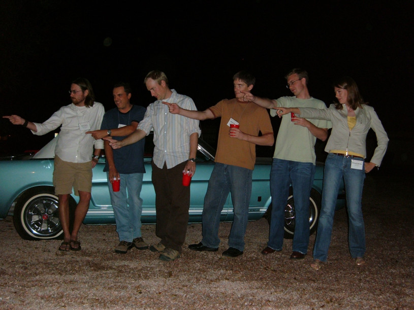 Six young people point to the right in front of a classic blue car