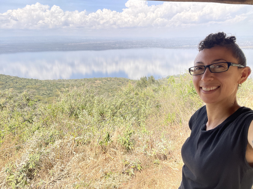 A woman in glasses smiles at the camera while taking a selfie in front of a green landscape