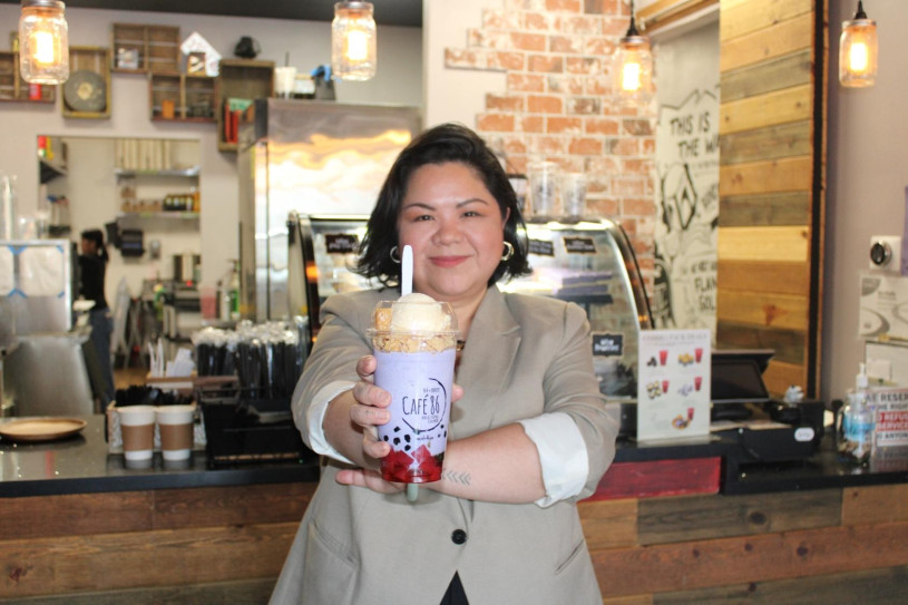 Filipino woman with short hair and a tan blazer holding out a purple halo-halo drink in front of a cafe counter.
