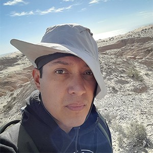 Head and shoulders portrait of Jose Alberto Cruz Silva wearing a hat with desert terrain and a blue sky with clouds in the background