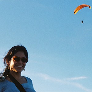 Head and shoulders portrait of Priscilla San Juan wearing sunglasses in the left foreground of the frame with a hang glider in the background in the top right of the frame