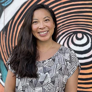 Whitney Tsai with long dark hair, wearing a black and white graphic-patterned shirt, against a background of orange, white, and blue-green background with concentric black circles