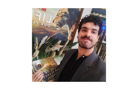 Head and shoulders portrait of Dr. Sergio Martínez Nebreda holding a book in front of a poster of a saber-toothed cat