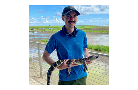 Zach Morris holding a gator with water and a cloudy blue sky in the background